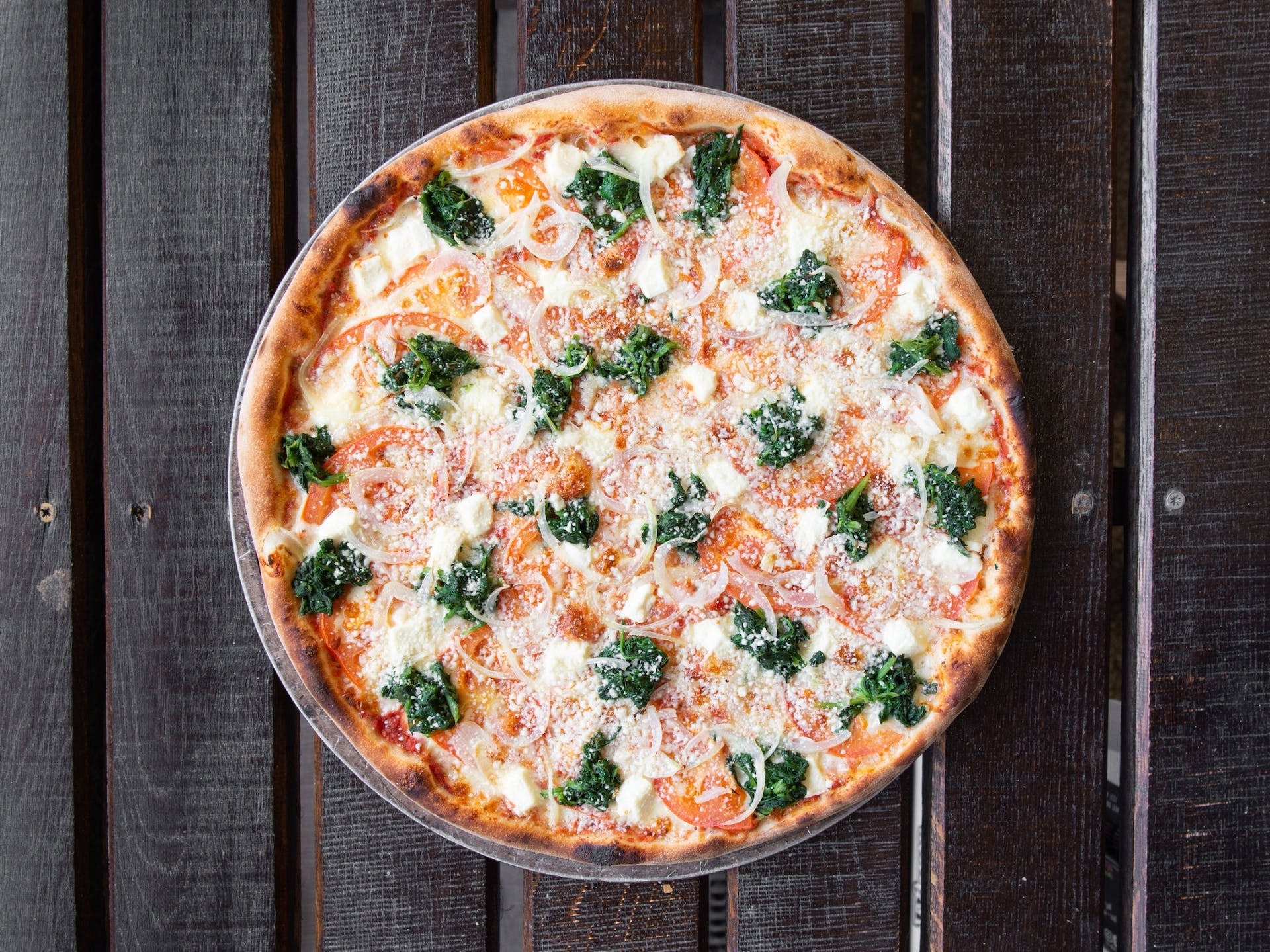 Pizza ze szpinakiem, pomidorami, cebulą i serem, posypana parmezanem, na drewnianym stole z ciemnych desek.