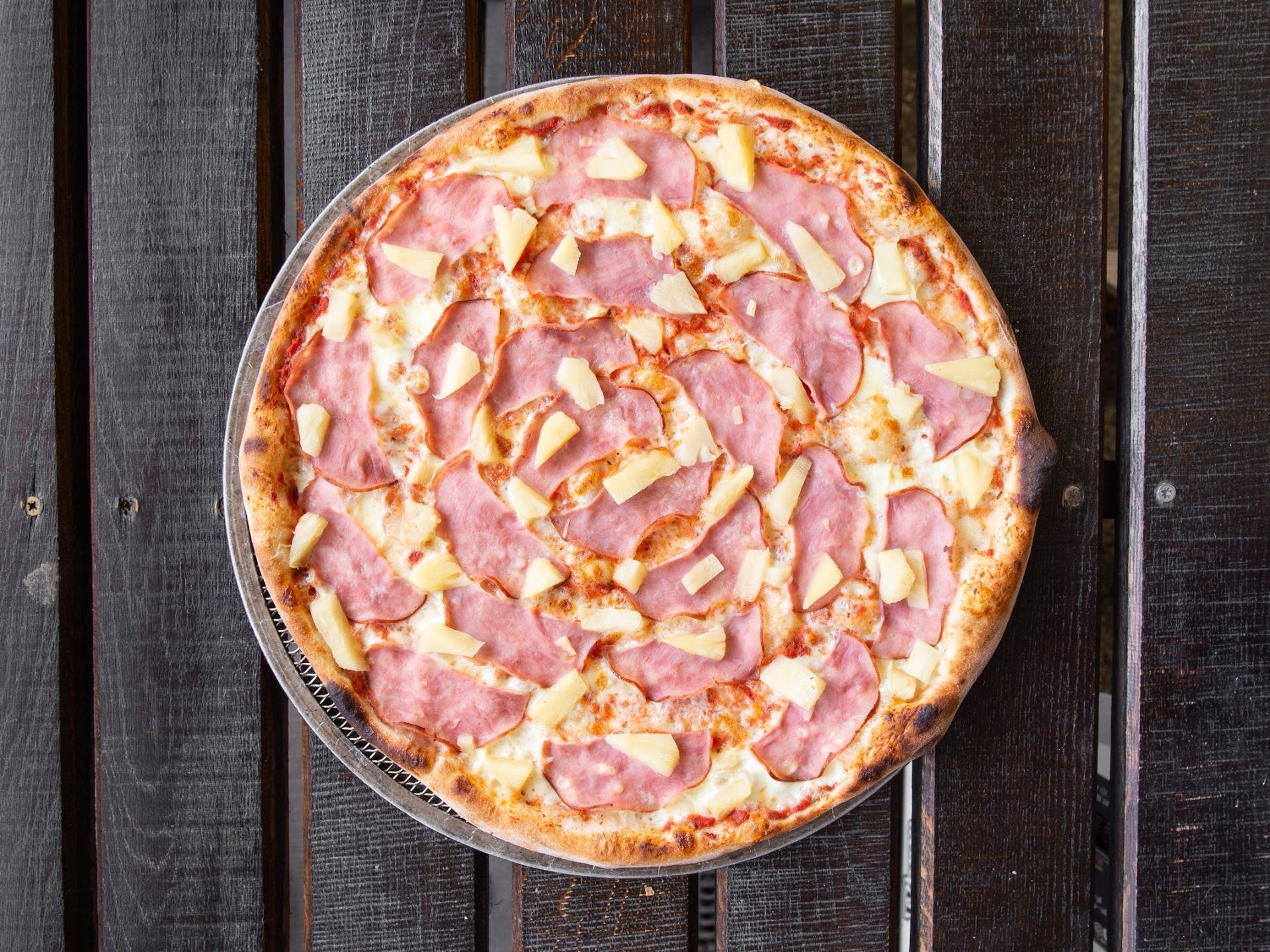 Pizza hawajska z szynką i ananasem na metalowej tacy, ułożona na ciemnym, drewnianym stole z widoczną strukturą drewna.