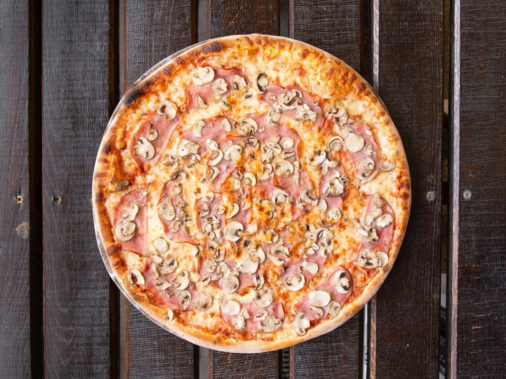 Pizza z szynką i pieczarkami na ciemnym, drewnianym stole z widocznymi śrubami.