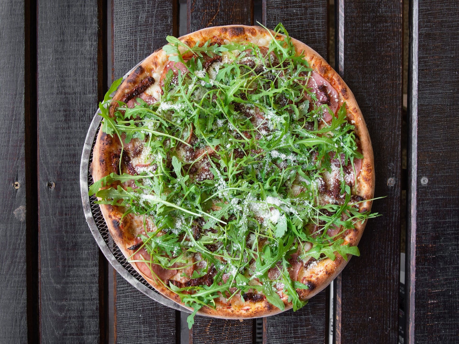 Pizza z szynką parmeńską, suszonymi pomidorami i rukolą posypana parmezanem, serwowana na metalowej siatce na tle ciemnego drewnianego stołu.