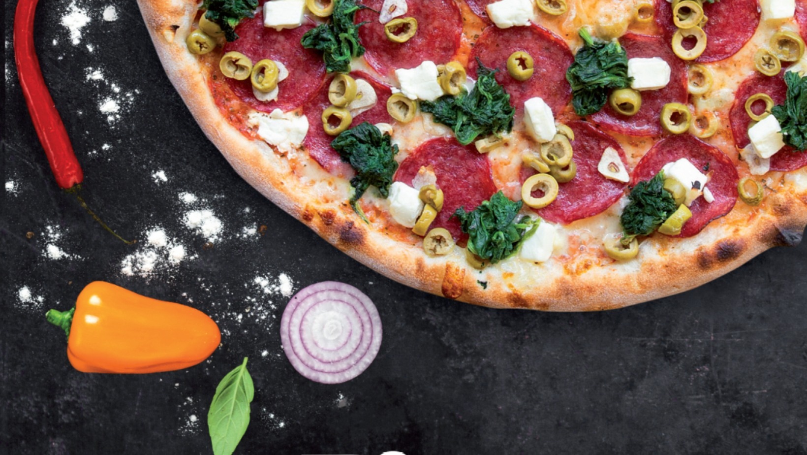 Pizza z salami, szpinakiem, oliwkami i serem feta na ciemnym tle z papryczką chili, małą żółtą papryką, plasterkiem czerwonej cebuli i listkiem bazylii obok.