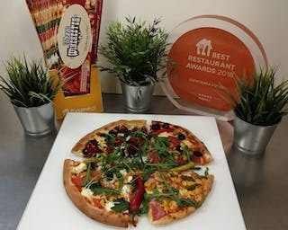 Pizza podzielona na cztery różne części, każda z innym rodzajem dodatków, ułożona na białym kwadratowym talerzu. W tle widoczne trzy małe roślinki w srebrnych doniczkach, opakowanie produktu...