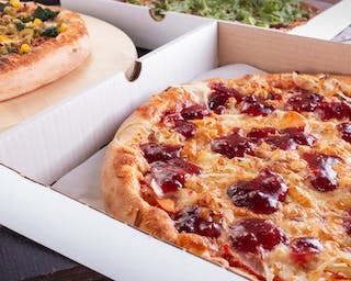 Pizza z dżemem wiśniowym w białym kartonie, obok pizza z kukurydzą i szpinakiem na drewnianej desce, widoczne inne pudełka z pizzą w tle.