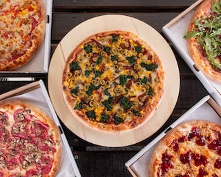 Cztery pizze w kartonach oraz na drewnianej desce do pizzy. Jedna pizza z kukurydzą i szpinakiem na środku kadru.