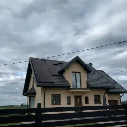 Dom jednorodzinny z zainstalowanymi panelami słonecznymi na dachu, widok zza czarnego ogrodzenia, pochmurne niebo.
