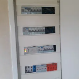 Otwarta skrzynka elektryczna z widocznymi bezpiecznikami, przewodami i modułami zabezpieczającymi, zainstalowana w pomieszczeniu z jasnymi ścianami.