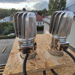 Dwa błyszczące, metalowe kominki wentylacyjne z turbiną na dachu z papy, zamontowane na płycie OSB, z widocznymi przewodami wentylacyjnymi i elementami konstrukcji dachu w tle zabudowa jednorodzinna.