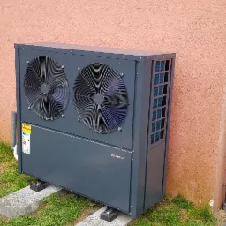 Szara pompa ciepła typu powietrze-woda, model Krommler, zamontowana na betonowych podstawach przy ścianie domu z rynną spustową, widok z poziomu trawnika.