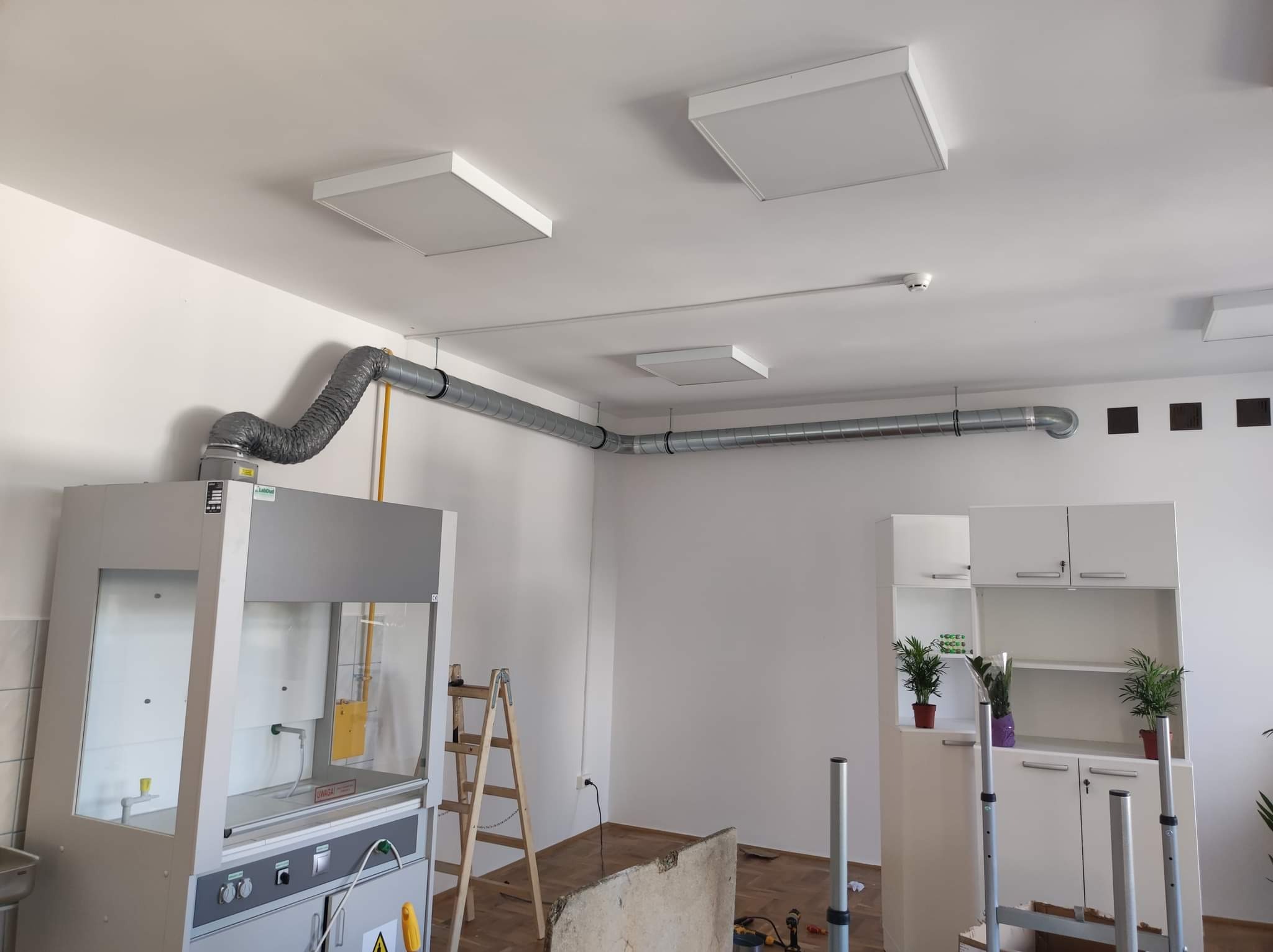 Wnętrze laboratorium z zainstalowanym dygestorium z podłączonym przewodem wentylacyjnym, widoczne również panele sufitowe i drabina.