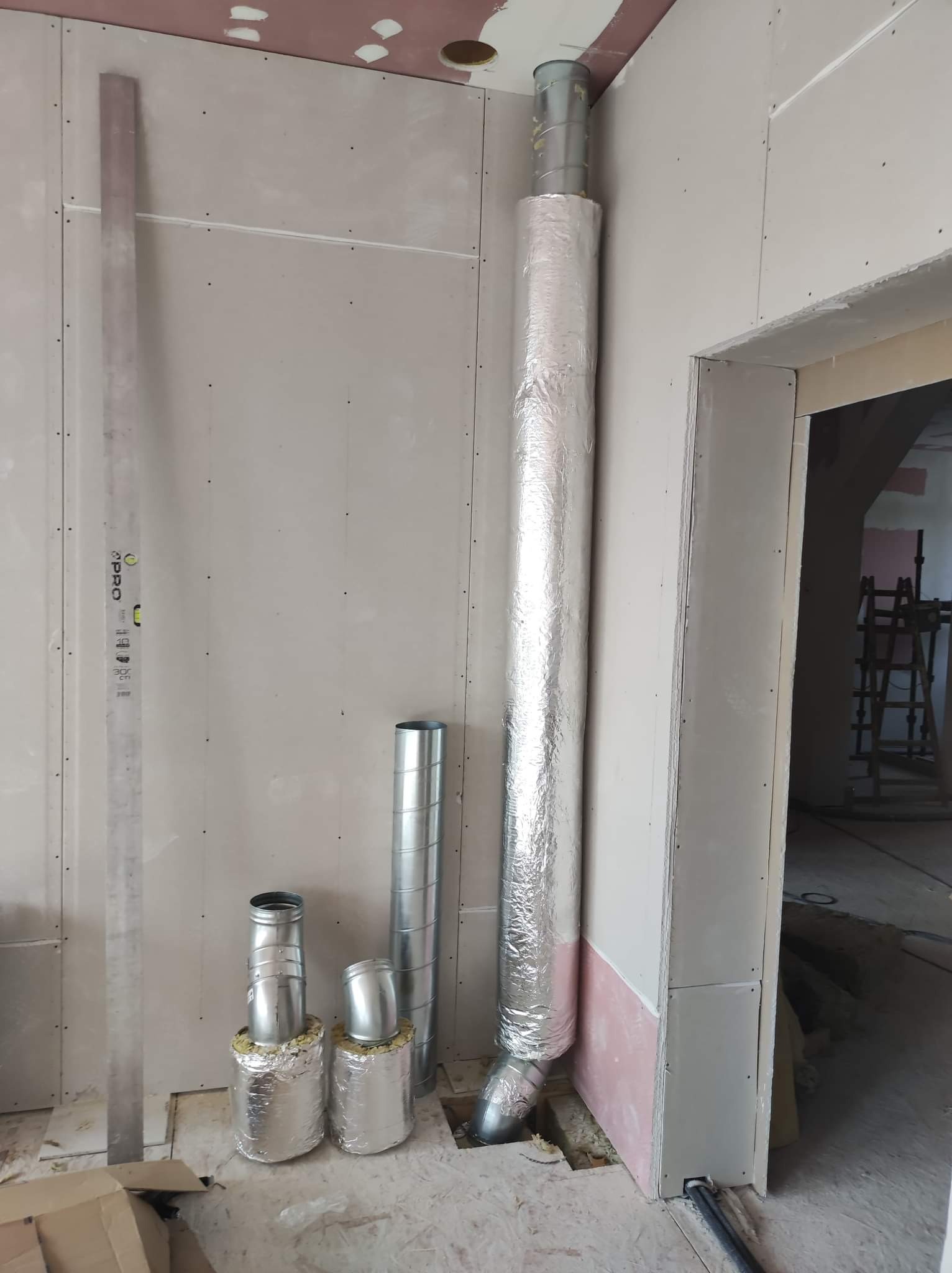 Wnętrze w trakcie wykańczania z widocznymi elementami instalacji wentylacyjnej: pionowy, izolowany przewód wentylacyjny, odcinki rur i poziomica oparta o ścianę z płyt gipsowo-kartonowych.