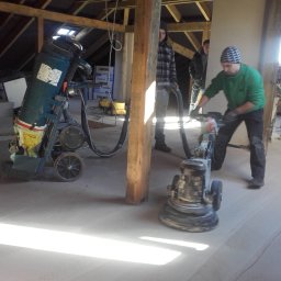 Usługi Budowlane - Inwestycyjne - Pracownik w zielonej bluzie szlifuje posadzkę na poddaszu przy użyciu maszyny do szlifowania, obok stoi odkurzacz przemysłowy, widoczne drewniane elementy konstrukcyjne dachu i okno dachowe.