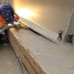 Usługi Budowlane - Inwestycyjne - Pracownik w ubraniu roboczym, klęcząc, rozprowadza szary płynny materiał na betonowej powierzchni za pomocą węża; widoczne ślady zabrudzeń i niedoskonałości betonu.