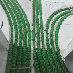 Usługi Budowlane - Inwestycyjne - Zielone rury karbowane ułożone w posadzce i zamocowane metalowymi taśmami, wypełnione pianką montażową, widoczne czarne przewody.