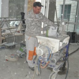 Usługi Budowlane - Inwestycyjne - Pracownik budowlany dosypuje worek zaprawy do maszyny do wylewania posadzek w surowym wnętrzu budynku w trakcie prac wykończeniowych.