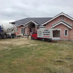 Betonomieszarka Agilia i samochód dostawczy z napisem 'Wylewki anhydrytowe z gwarancją' na tle domu w trakcie budowy, widoczny pracownik w pomarańczowej kamizelce.