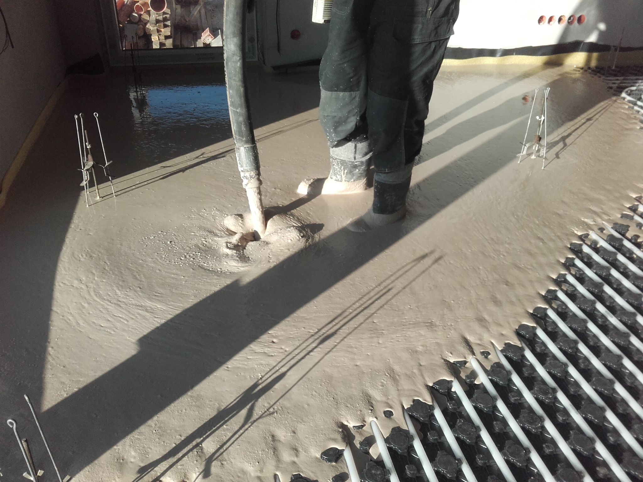 Wylewanie płynnej masy na ogrzewanie podłogowe, widoczne rury instalacji i pracownik w ubrudzonych butach roboczych, cień węża do wylewania.