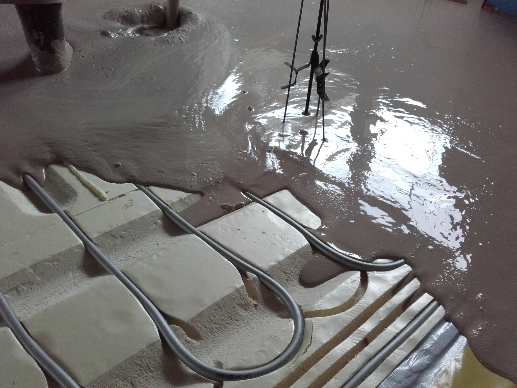 Wylewanie płynnej masy na podkład z rurami ogrzewania podłogowego, widoczny mikser zanurzony w masie.