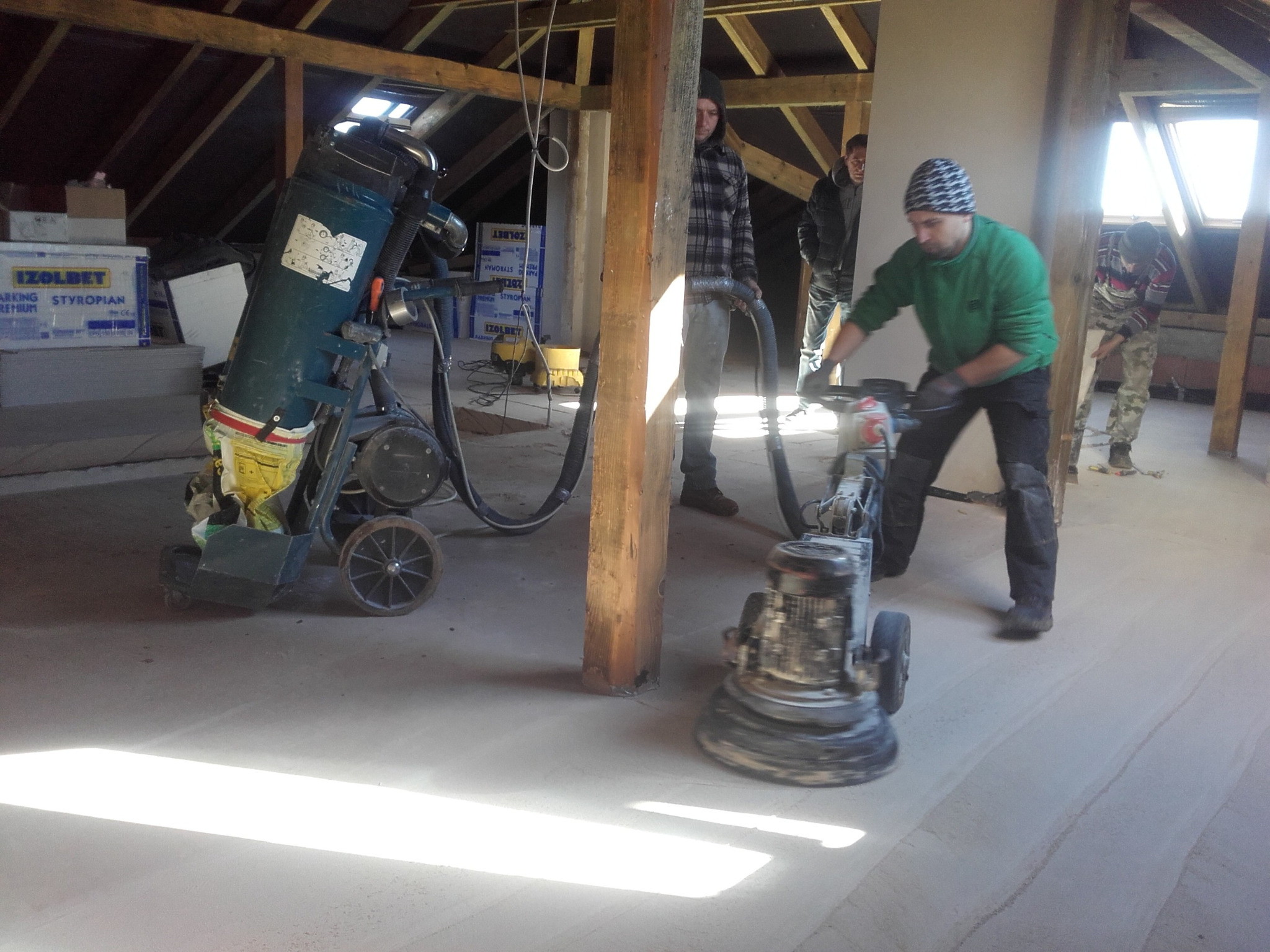 Pracownik w zielonej bluzie szlifuje posadzkę na poddaszu przy użyciu maszyny do szlifowania, obok stoi odkurzacz przemysłowy, widoczne drewniane elementy konstrukcyjne dachu i okno dachowe.