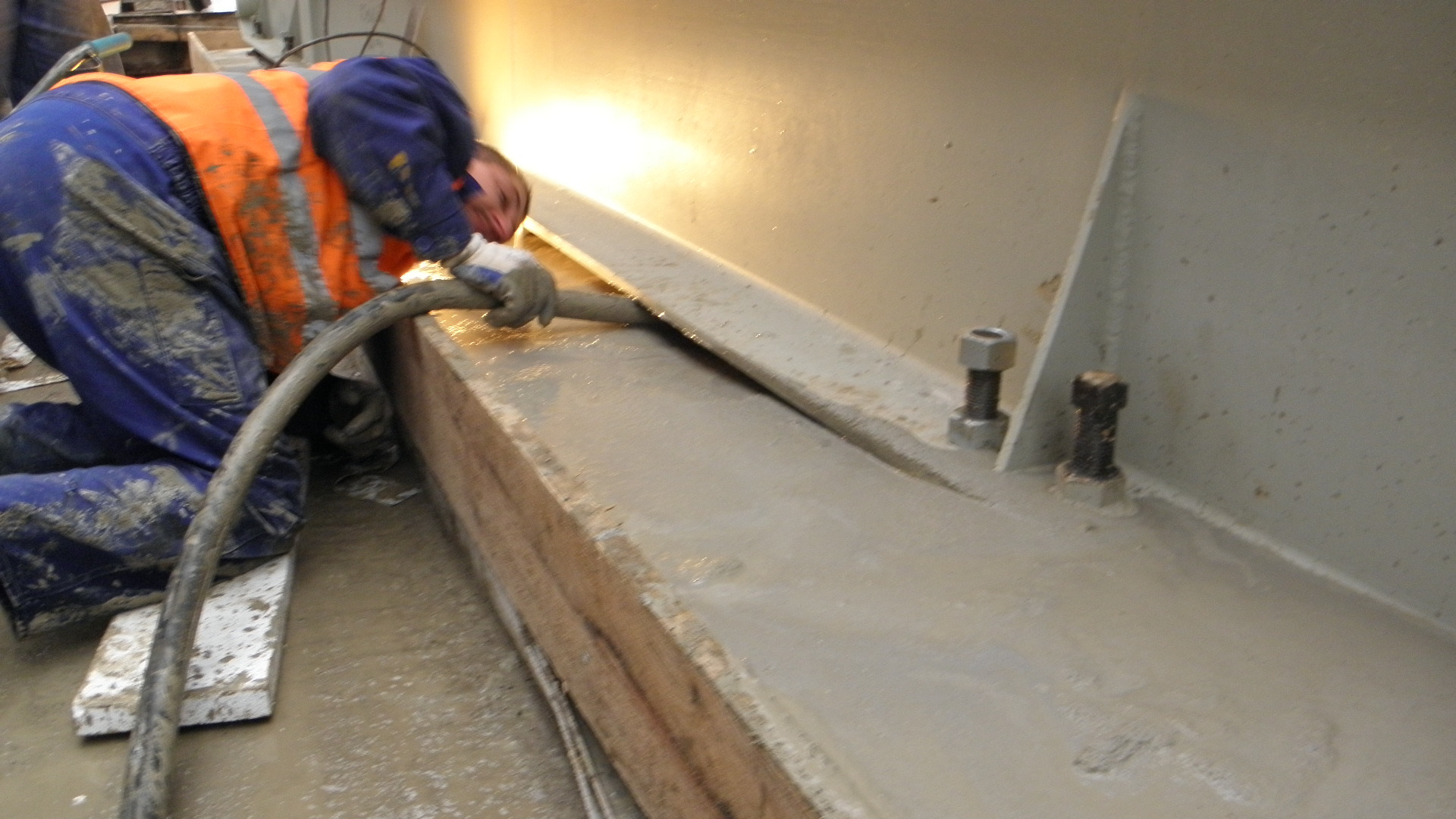 Pracownik w ubraniu roboczym, klęcząc, rozprowadza szary płynny materiał na betonowej powierzchni za pomocą węża; widoczne ślady zabrudzeń i niedoskonałości betonu.