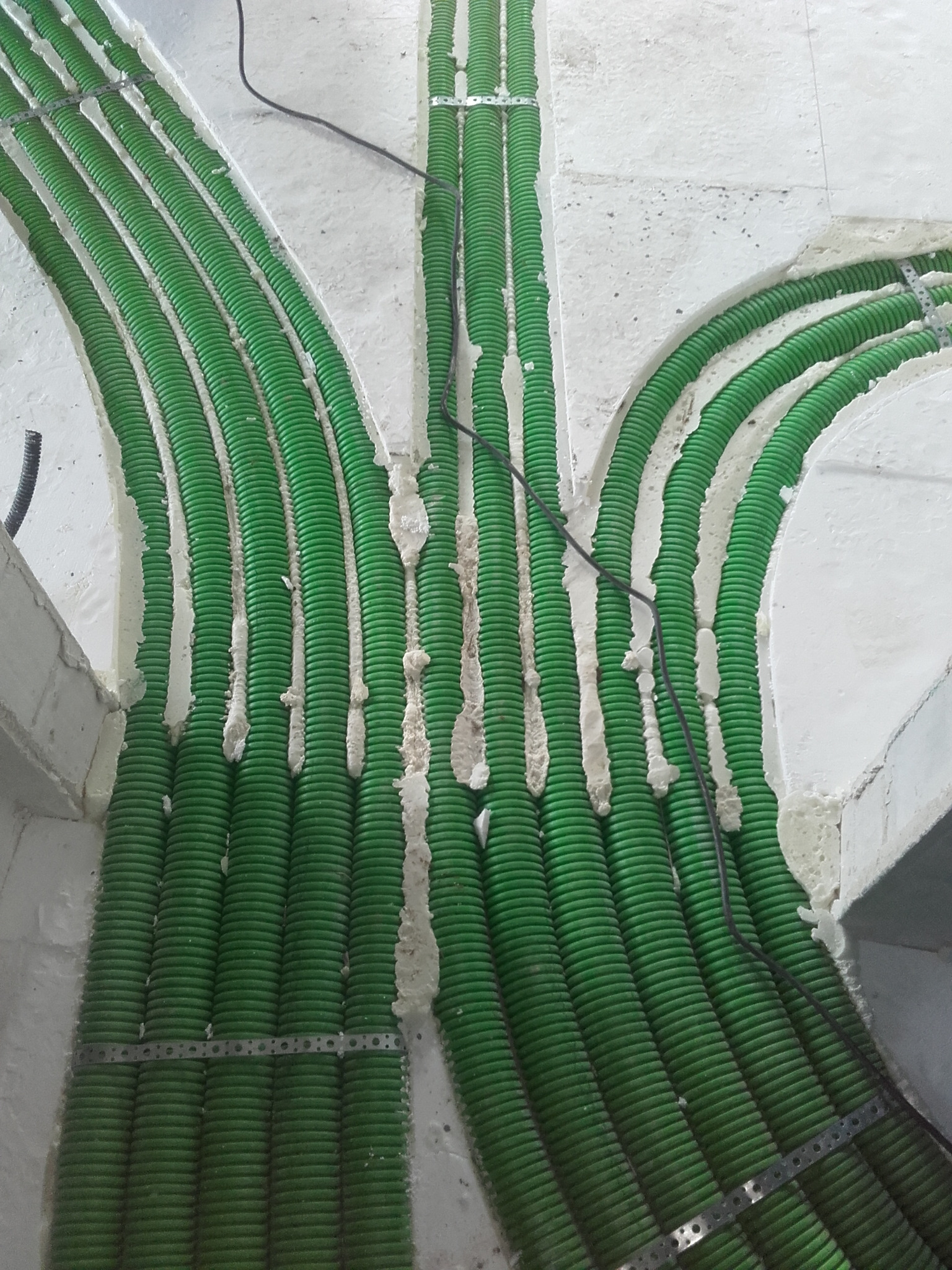 Zielone rury karbowane ułożone w posadzce i zamocowane metalowymi taśmami, wypełnione pianką montażową, widoczne czarne przewody.