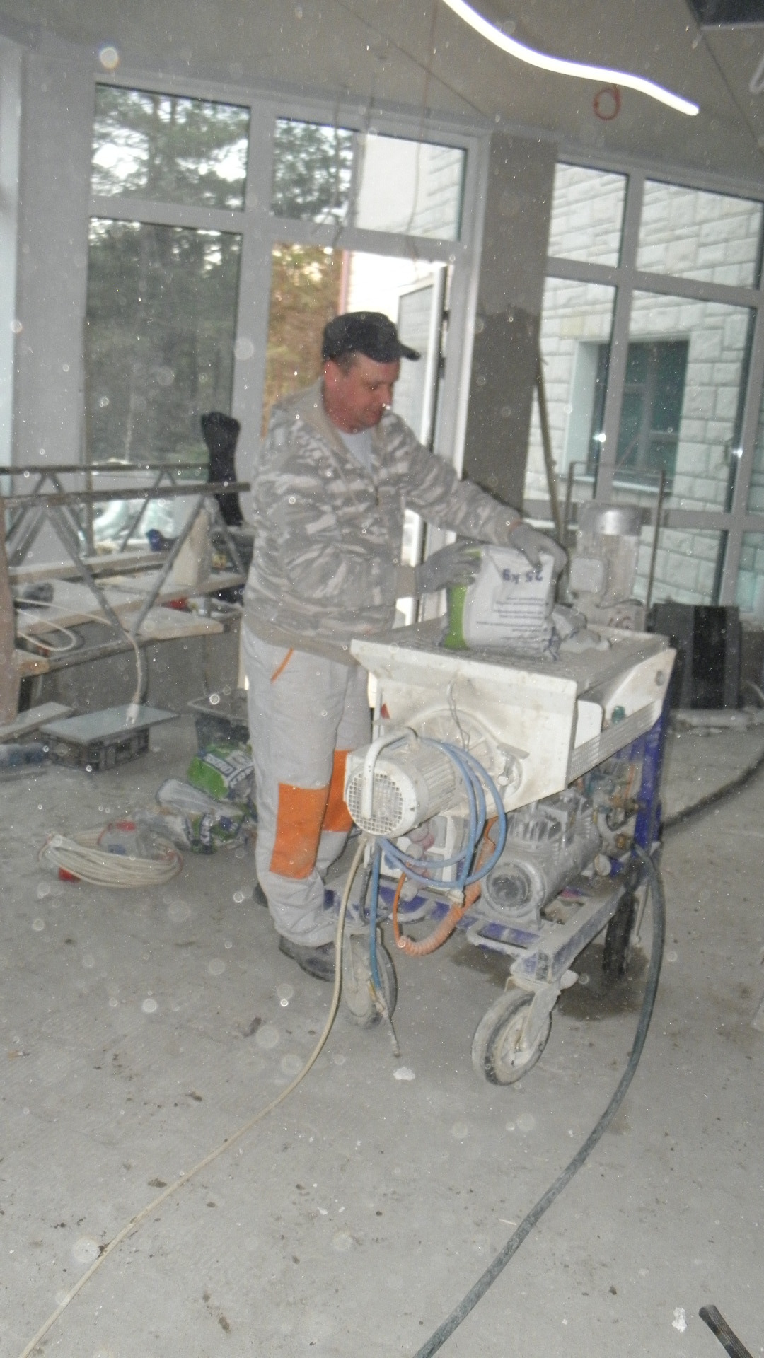 Pracownik budowlany dosypuje worek zaprawy do maszyny do wylewania posadzek w surowym wnętrzu budynku w trakcie prac wykończeniowych.