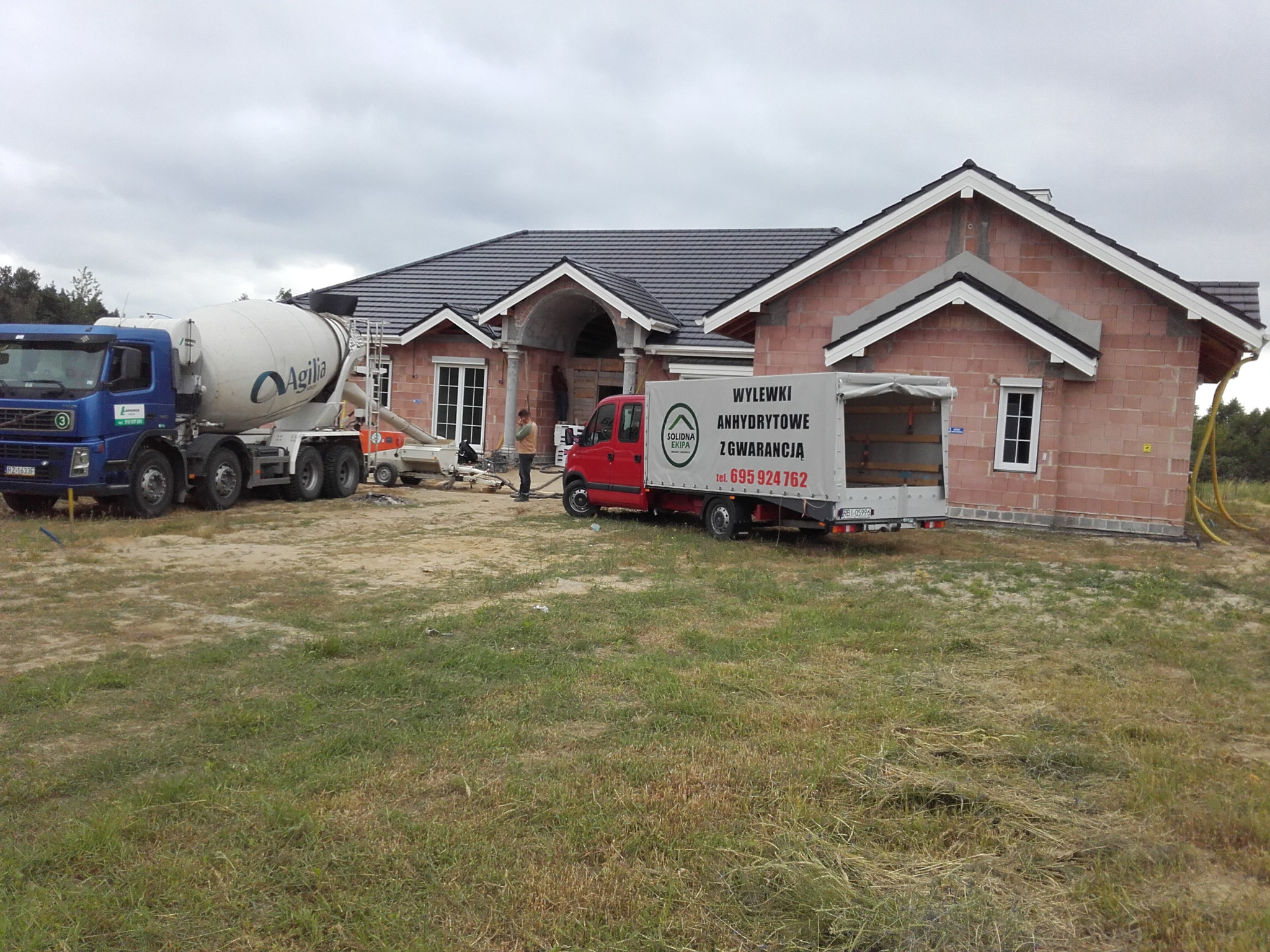 Betonomieszarka Agilia i samochód dostawczy z napisem 'Wylewki anhydrytowe z gwarancją' na tle domu w trakcie budowy, widoczny pracownik w pomarańczowej kamizelce.