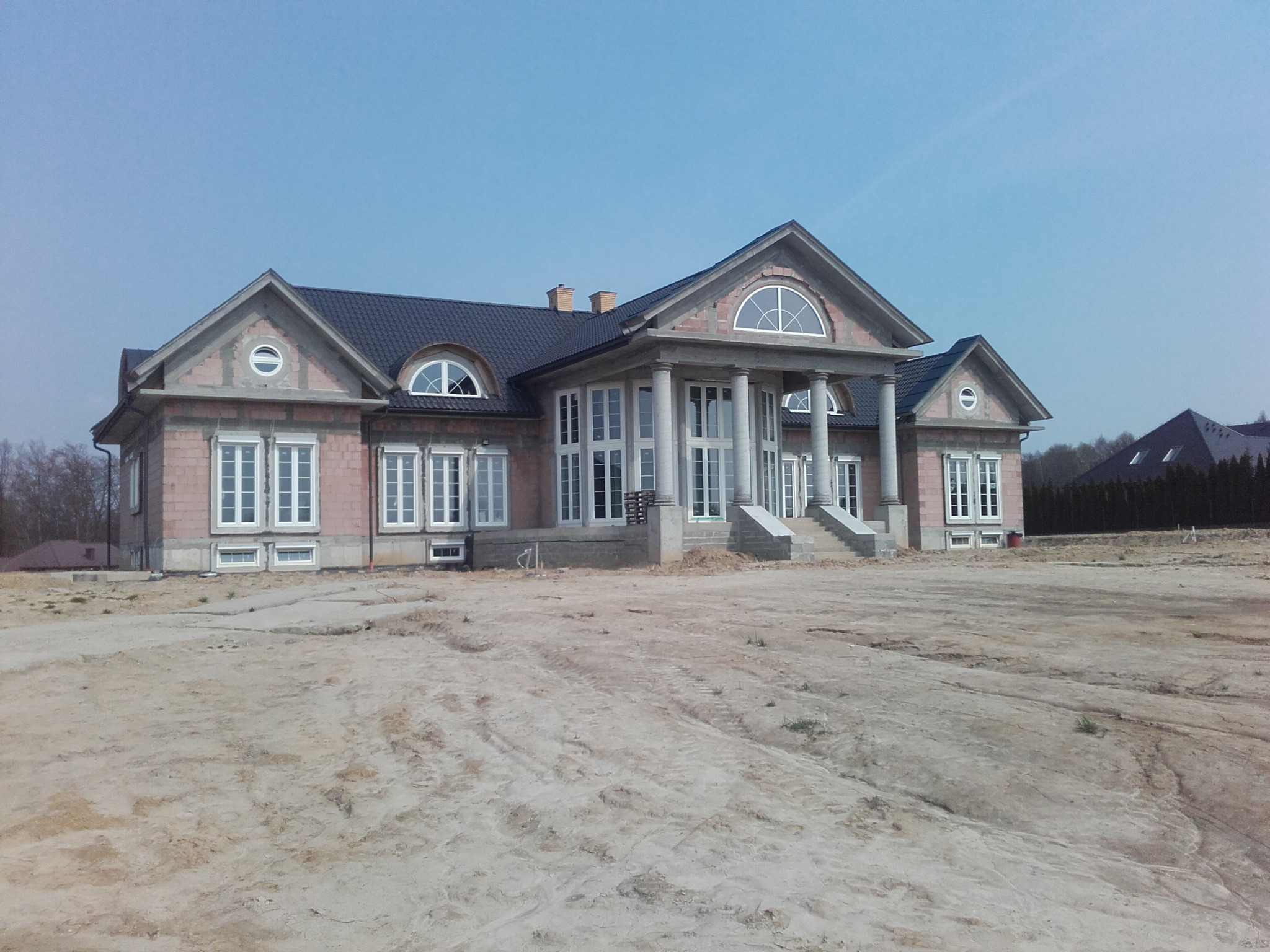 Niedokończony dom z cegły z kolumnami i licznymi oknami w stylu klasycystycznym, otoczony piaszczystym terenem i widocznym zarysem sąsiedniego budynku.