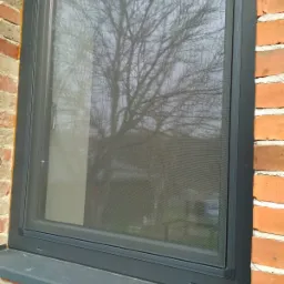 Zamknięte okno z szarą ramą, z zamontowaną moskitierą, widok na drzewo bez liści za oknem, mur z czerwonej cegły po bokach okna.