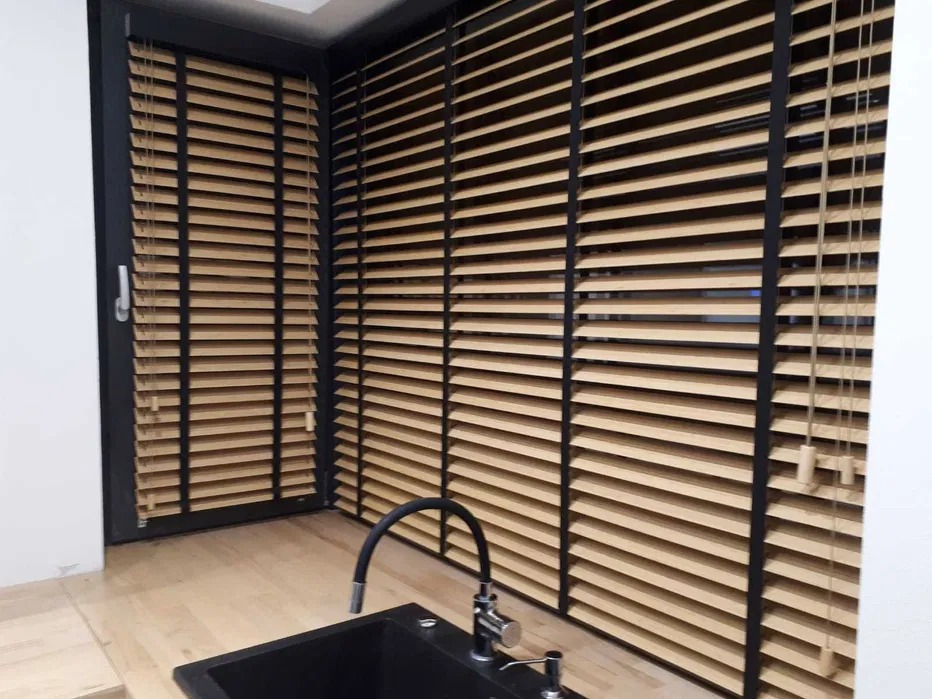 Jasne, drewniane żaluzje poziome z czarnymi ramami zamontowane w oknie i na ścianie, widoczny fragment zlewu kuchennego z czarnym kranem.