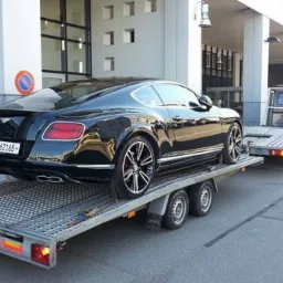 Czarny Bentley Continental GT na niskopodwoziowej przyczepie lawety, widok z tyłu, na tle nowoczesnego budynku z kolumnami.