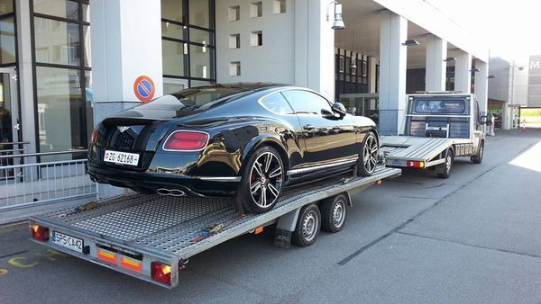 Czarny Bentley Continental GT na niskopodwoziowej przyczepie lawety, widok z tyłu, na tle nowoczesnego budynku z kolumnami.
