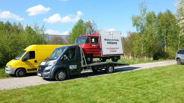 Laweta przewozi czerwony pojazd strażacki z reklamą na przyczepie, obok stoi żółty bus, na tle zieleni i błękitnego nieba.