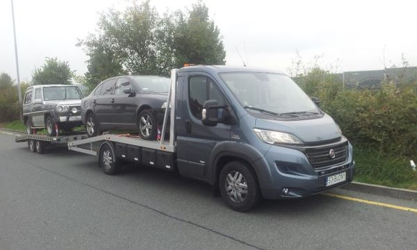 Niebieska laweta z dwoma samochodami: szarym SUV-em i czarną Skodą Octavią, zaparkowana na poboczu drogi w pochmurny dzień.