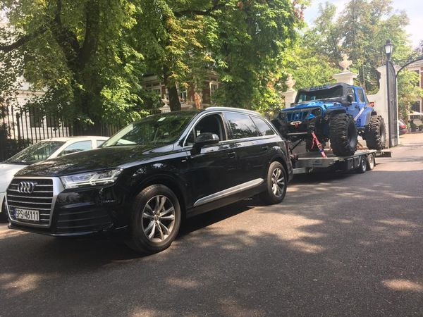 Czarny SUV Audi ciągnie na lawecie niebieskiego, terenowego Jeepa z bardzo dużymi kołami na tle drzew i bramy.