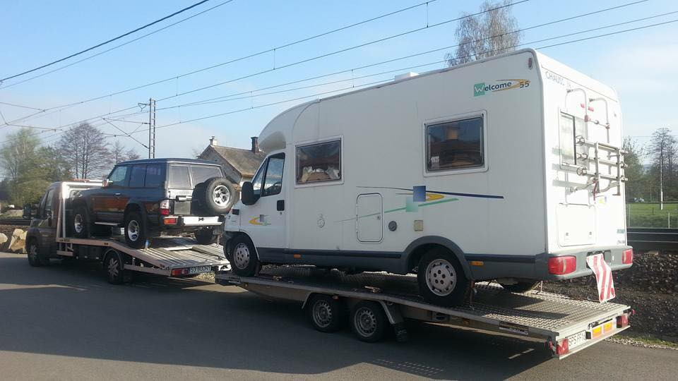 Laweta przewozi samochód terenowy i kamper marki Welcome 55 na tle torów kolejowych.
