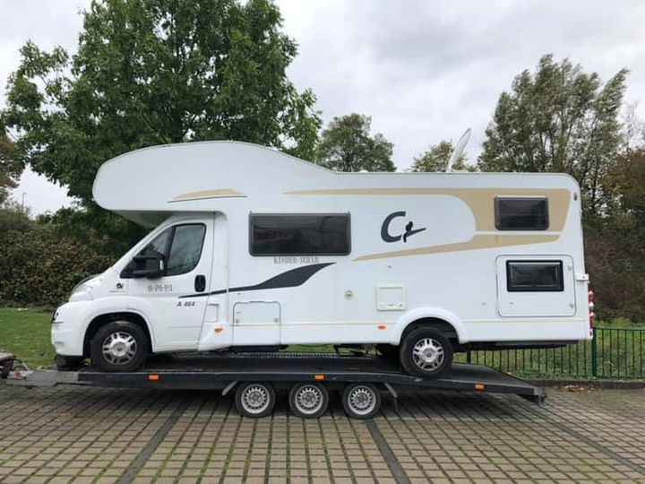 Biały kamper marki Ci, model Kinder-Schein, umieszczony na czarnej, niskopodwoziowej lawecie na tle drzew i szarej kostki brukowej.