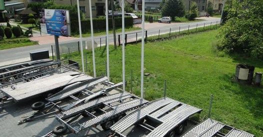 Rząd metalowych przyczep samochodowych na placu, widok z góry, w tle budynki i billboard reklamowy.