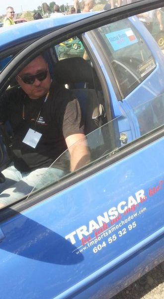 Mężczyzna w okularach przeciwsłonecznych siedzi w niebieskim samochodzie z logo firmy transportowej 'Transcar' na drzwiach.