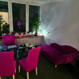 Gabinet psychoterapeutyczny z fioletowymi krzesłami, kanapą i szklanym stołem, oświetlony lampą, z roślinami doniczkowymi przy oknie i na szafce.