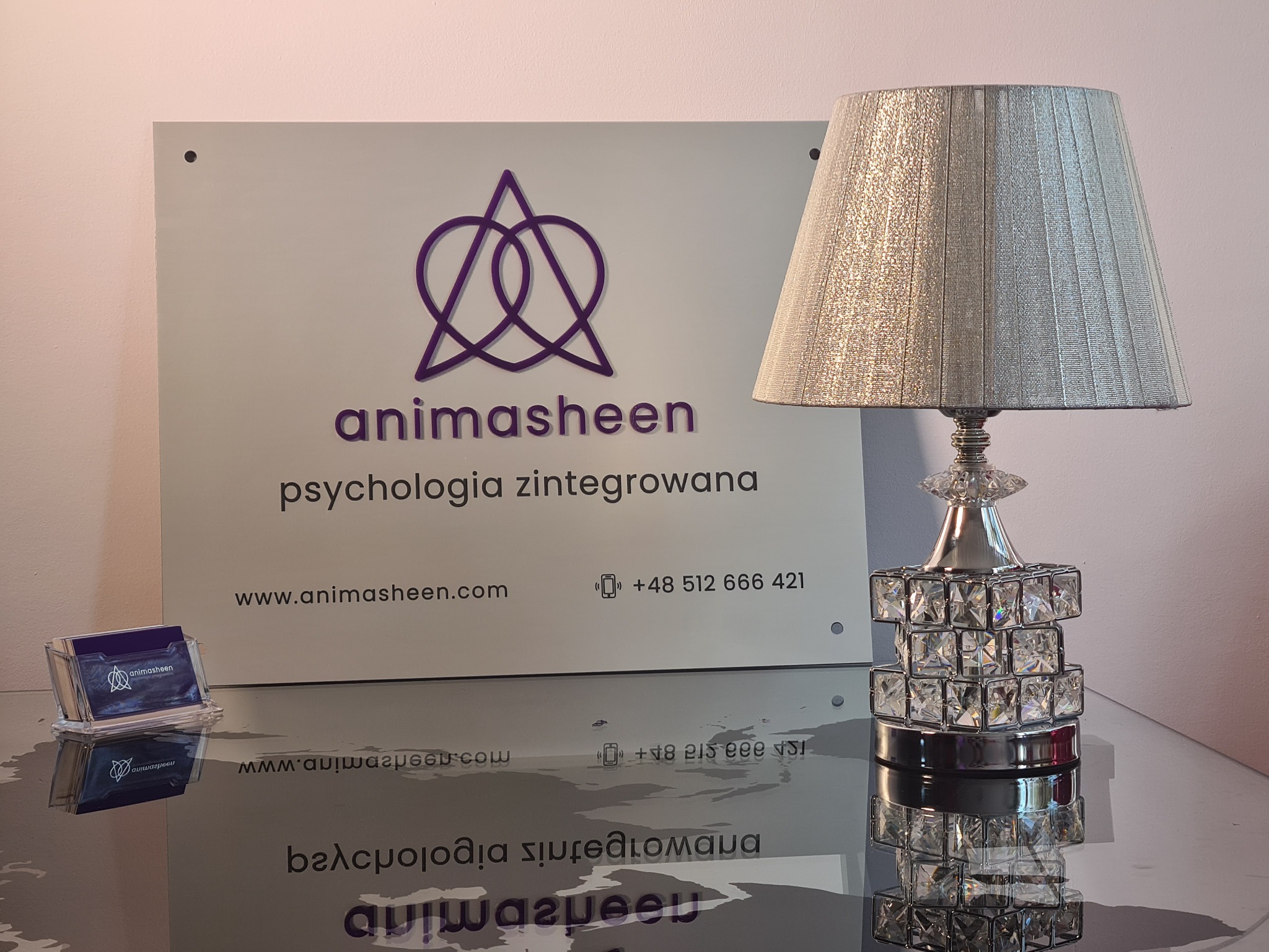 Tablica informacyjna z logo i napisem 'animasheen psychologia zintegrowana', wizytówki w przezroczystym pojemniku i ozdobna lampka na błyszczącej powierzchni.