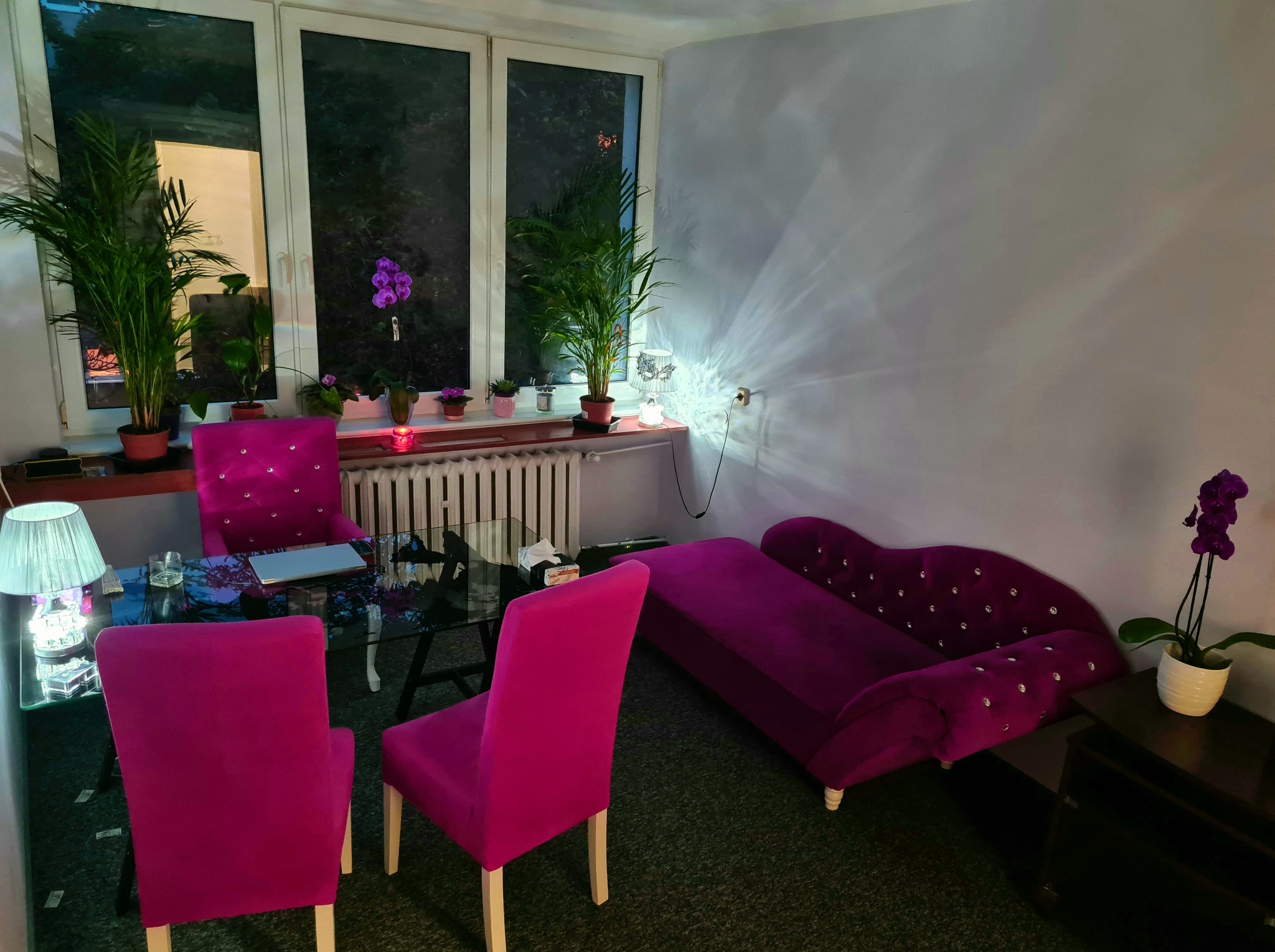 Gabinet psychoterapeutyczny z fioletowymi krzesłami, kanapą i szklanym stołem, oświetlony lampą, z roślinami doniczkowymi przy oknie i na szafce.
