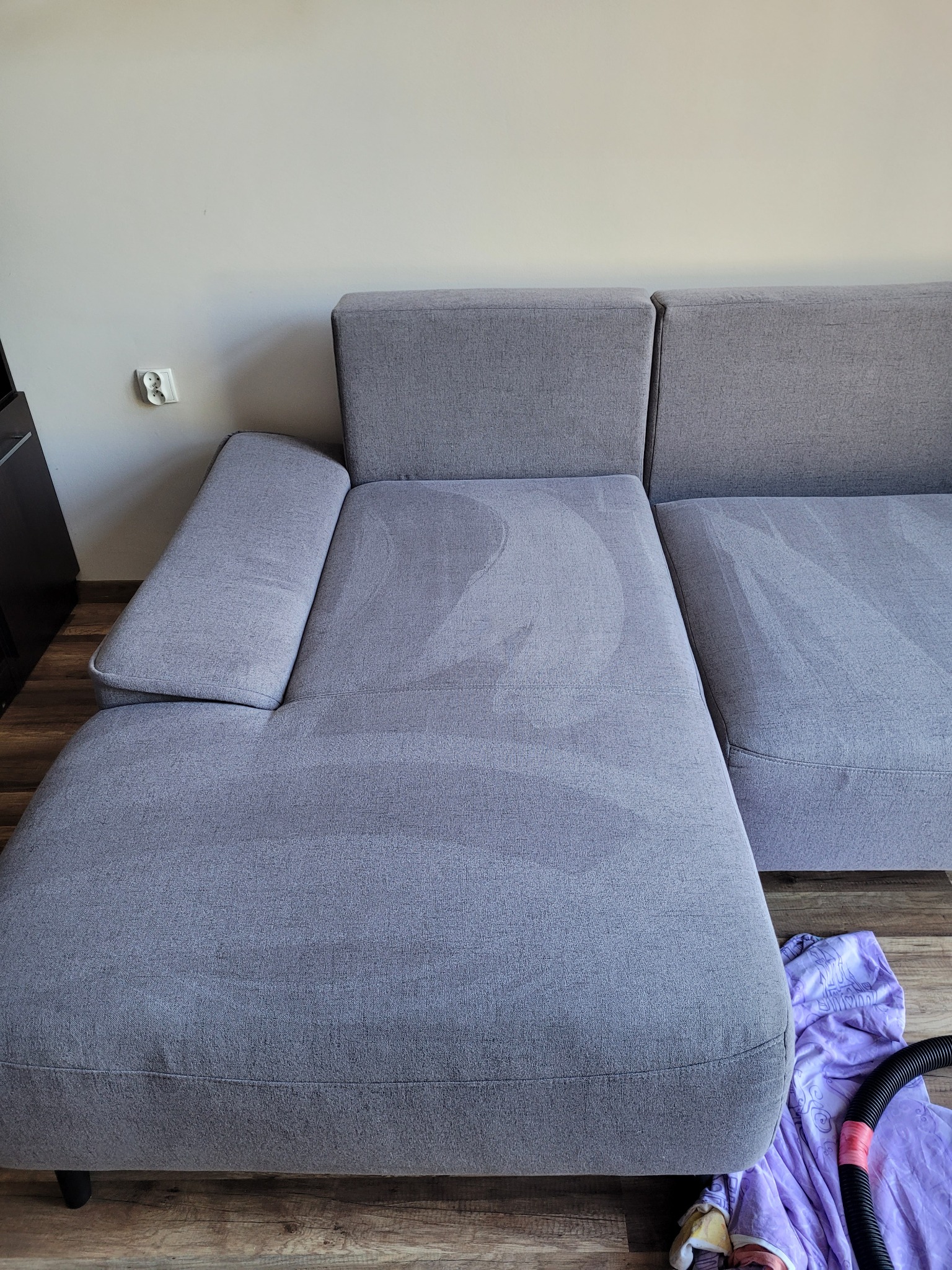 Szara, modułowa kanapa narożna z widocznymi śladami świeżo wypranej tapicerki, obok leży fioletowa ściereczka i wąż od odkurzacza piorącego.
