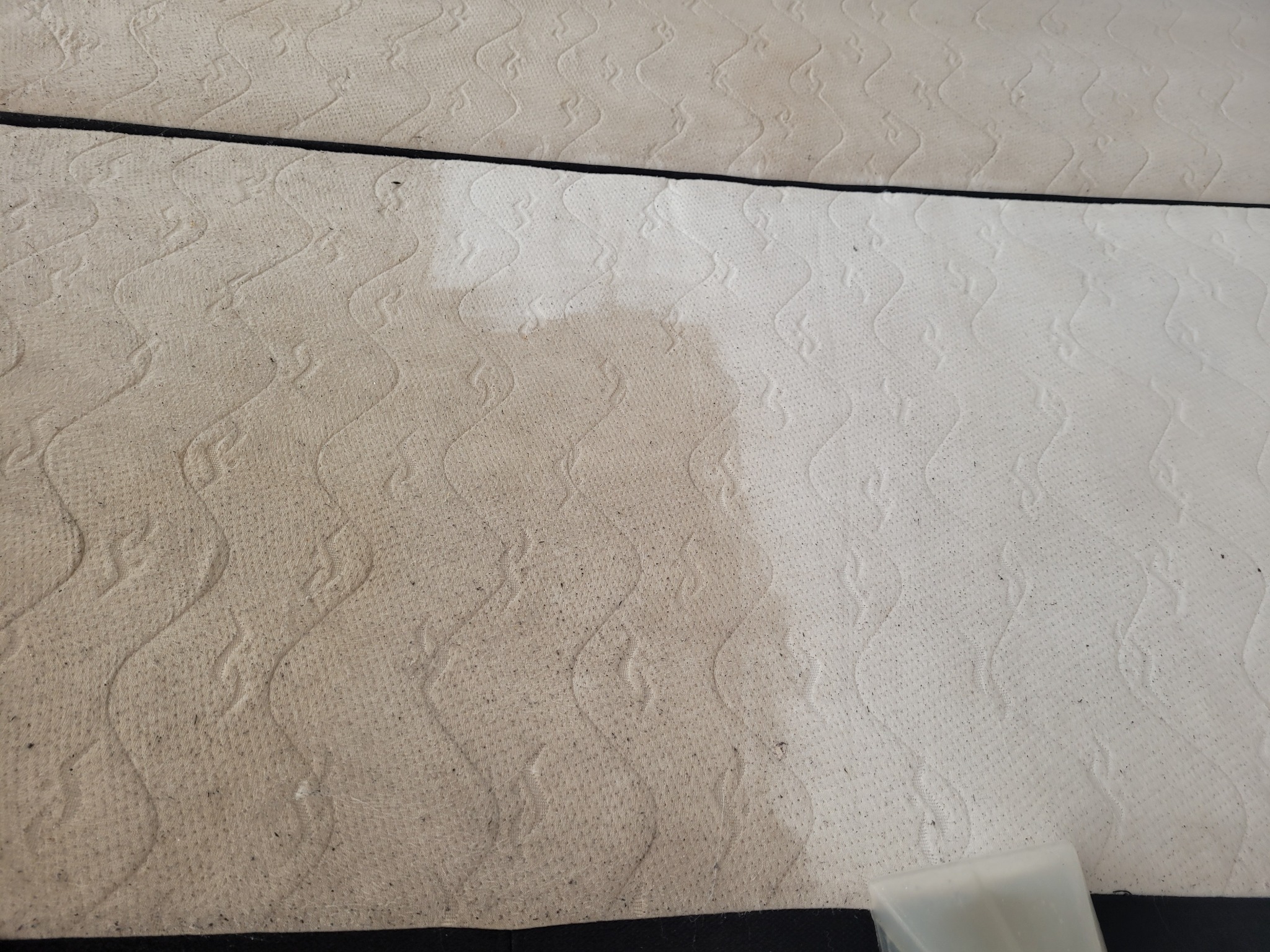 Częściowo wyczyszczony, jasny materac z wzorem, widoczne wyraźne różnice w kolorze i zabrudzeniach przed i po czyszczeniu, fragment dyszy czyszczącej w rogu.