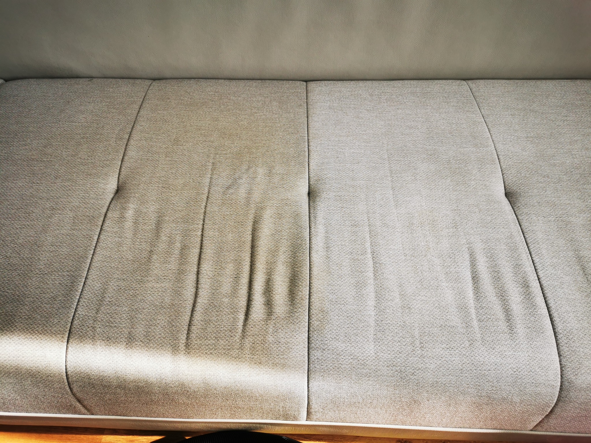 Widok z góry na fragment sofy z wyraźną różnicą w czystości: lewa strona wyraźnie brudniejsza, prawa świeżo wyczyszczona, z delikatnym cieniem padającym na czystą powierzchnię.