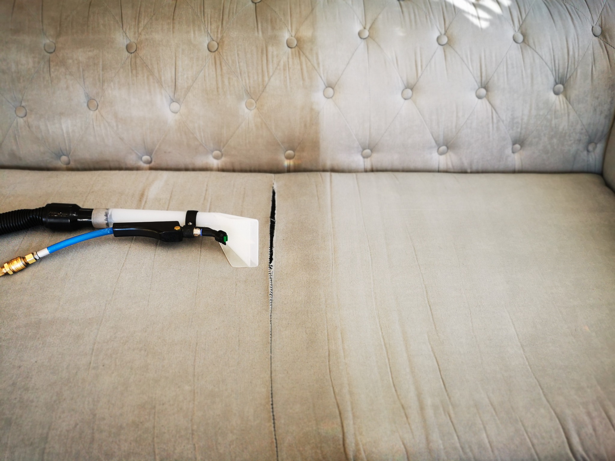 Czyszczenie tapicerowanej sofy z guzikami, widoczna różnica w odcieniu po prawej stronie, gdzie sofa jest czysta, oraz po lewej, gdzie jest jeszcze brudna, z dyszą czyszczącą i niebieskim wężem.