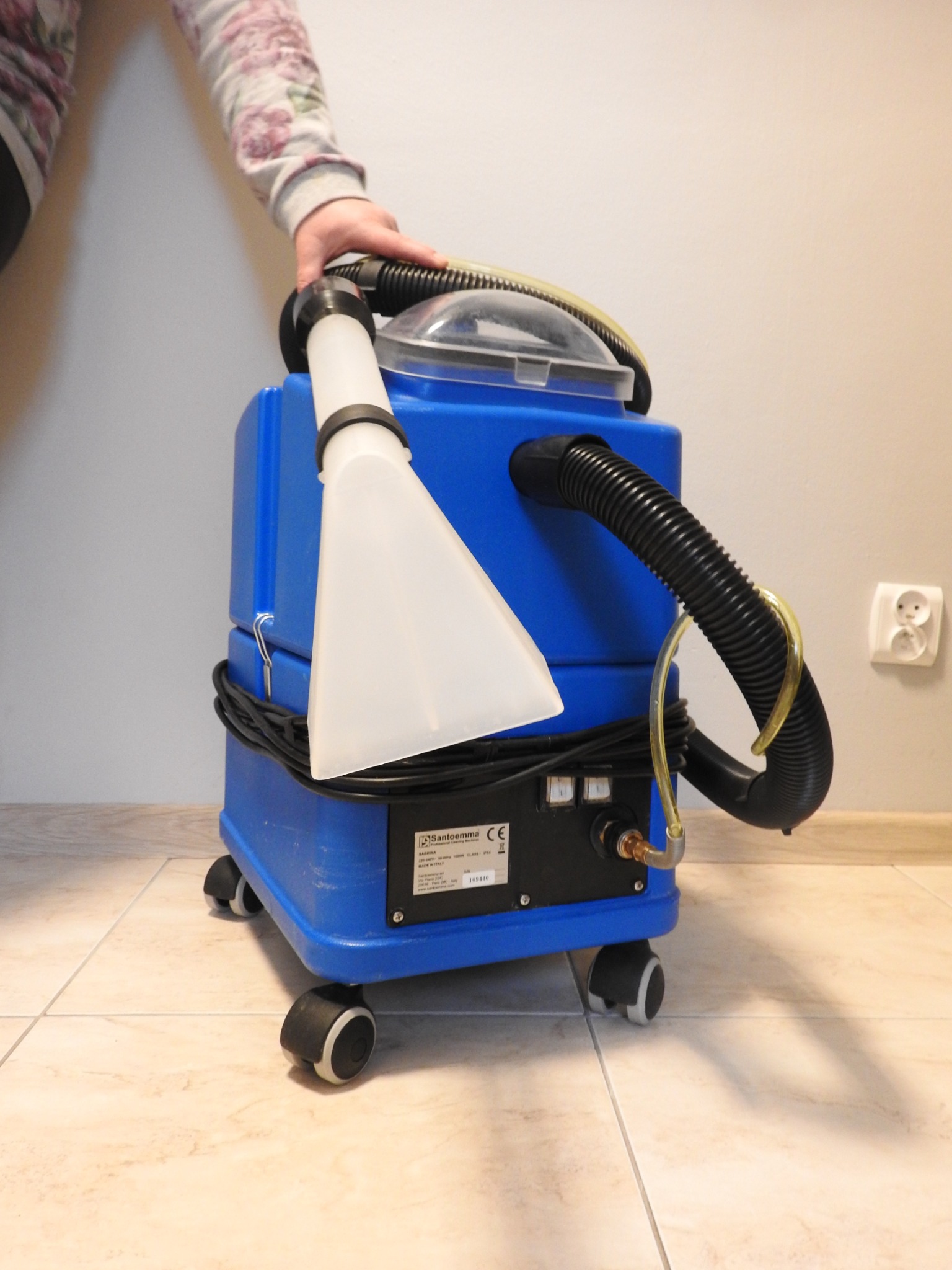 Niebieski odkurzacz piorący Santoemma na kółkach z białą ssawką i czarnym wężem, kabel owinięty wokół obudowy, fragment dłoni w kwiecistym rękawie na górze.