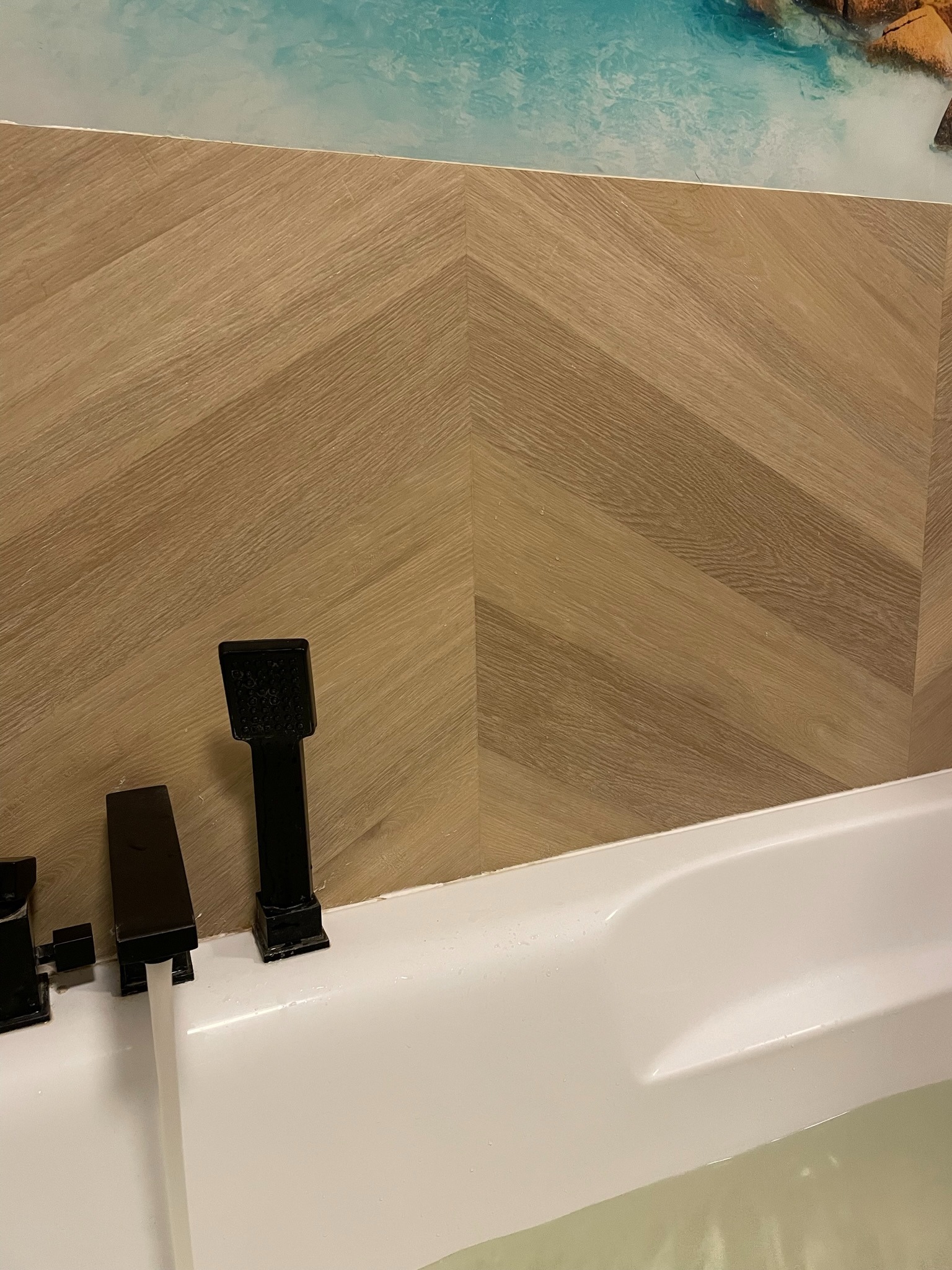 Biała wanna z czarną armaturą na tle ściany z drewnopodobnych paneli ułożonych w jodełkę, nad którymi widoczny fragment dekoracyjnej tapety z motywem morza.