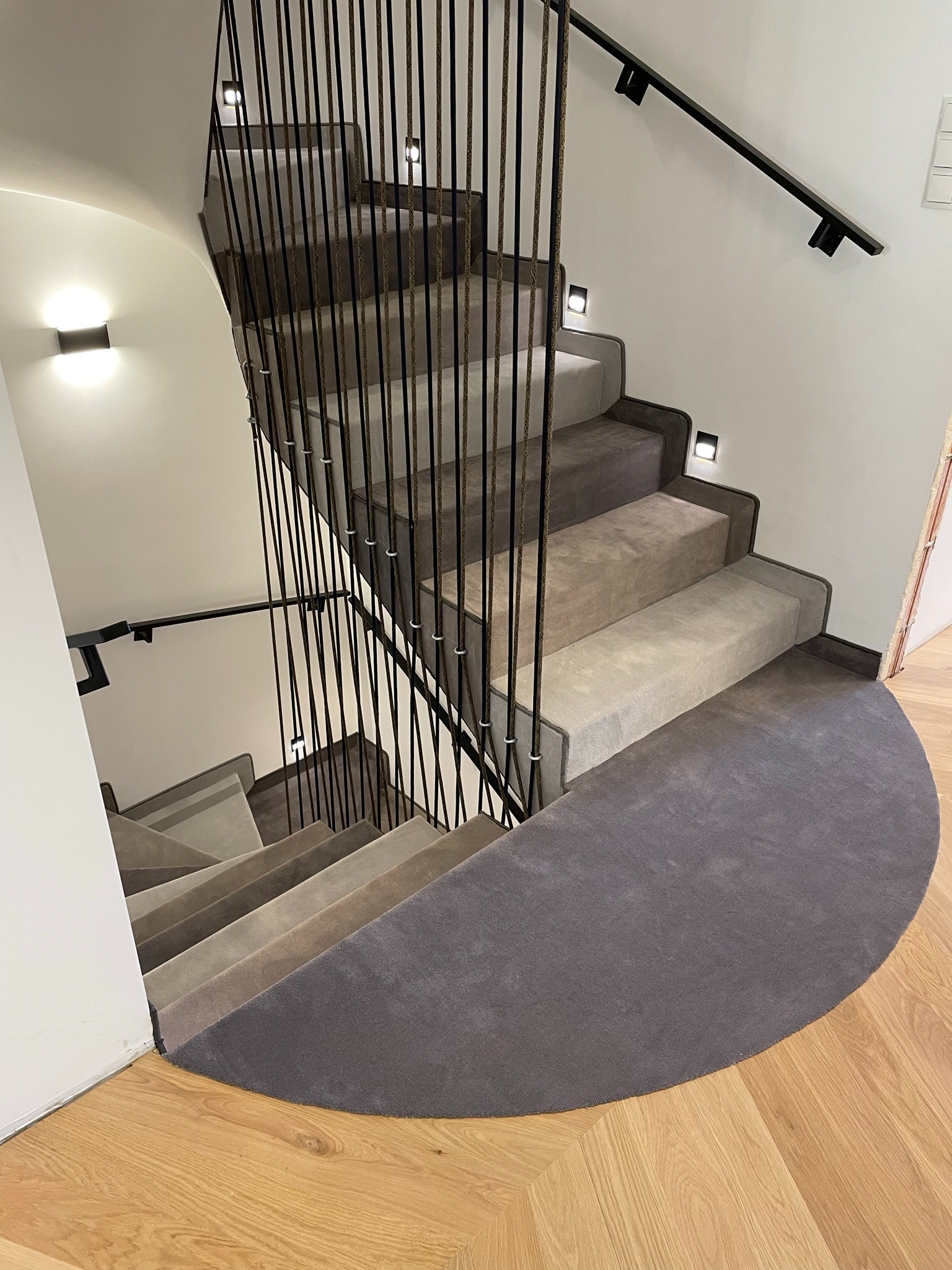 Wewnętrzne schody z wykładziną dywanową w odcieniach szarości, zabezpieczone czarną, metalową balustradą ze sznurkowym oplotem, oświetlone małymi, kwadratowymi lampami ściennymi.