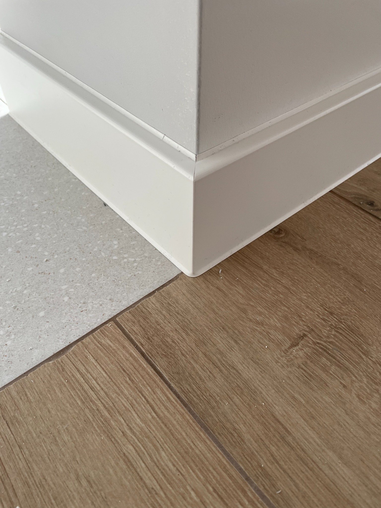 Detale wykończenia wnętrza: narożnik ściany z białą listwą przypodłogową, stykająca się z jasnoszarą terakotą i panelami podłogowymi imitującymi drewno.