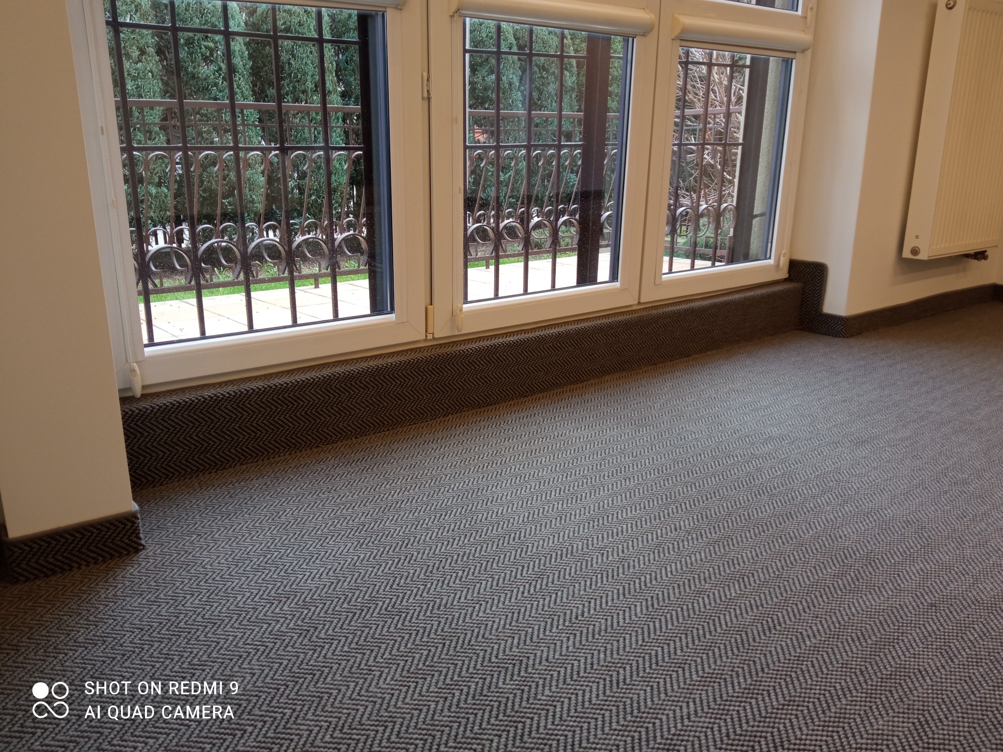 Wnętrze z nową wykładziną dywanową w jodełkę, widok na ozdobne okna z kratami i zieleń za nimi, grzejnik ścienny po prawej.